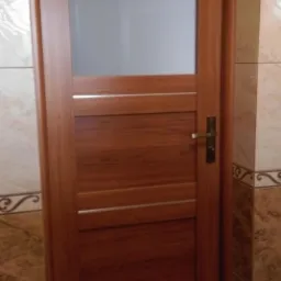 Zamknięte, brązowe drzwi wewnętrzne z poziomymi panelami i szybą, zamontowane w łazience z brązowymi płytkami.