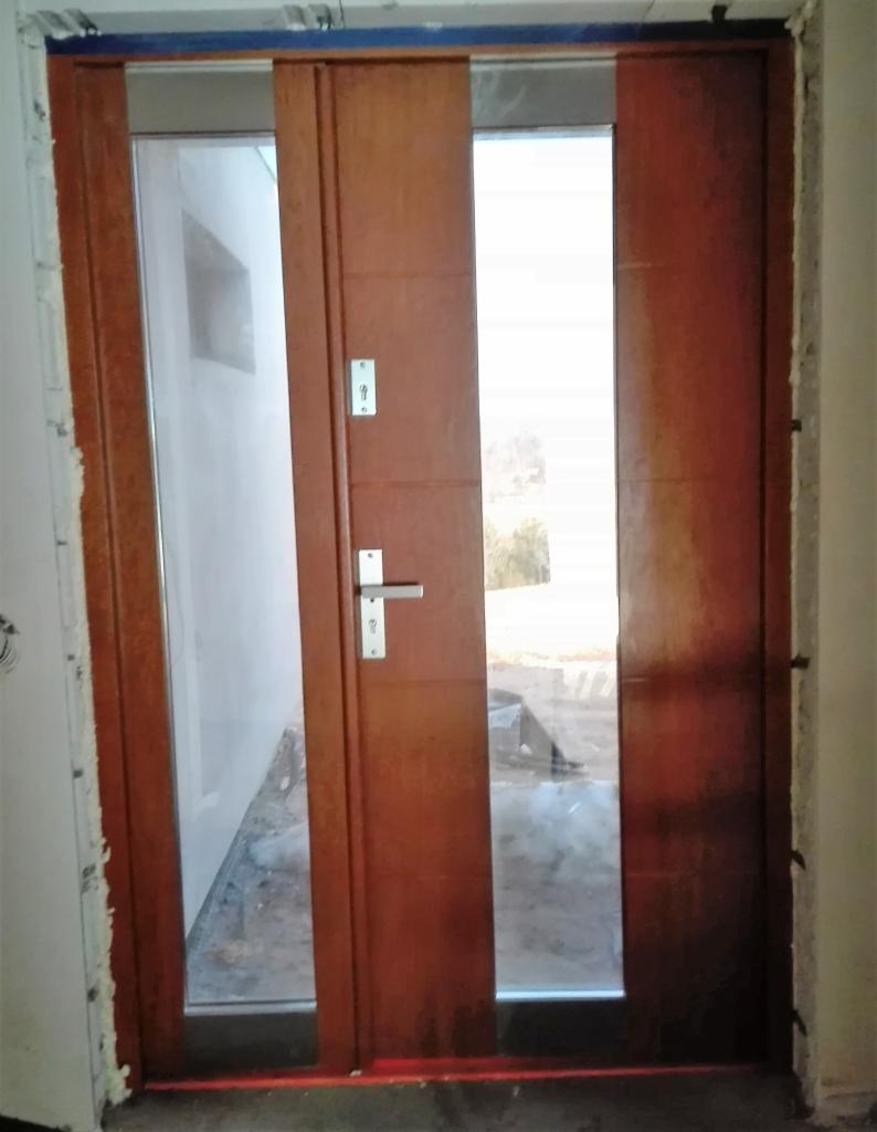 Brązowe drzwi wejściowe z przeszkleniem, widoczne ślady montażu, surowy stan wykończenia wnętrza.