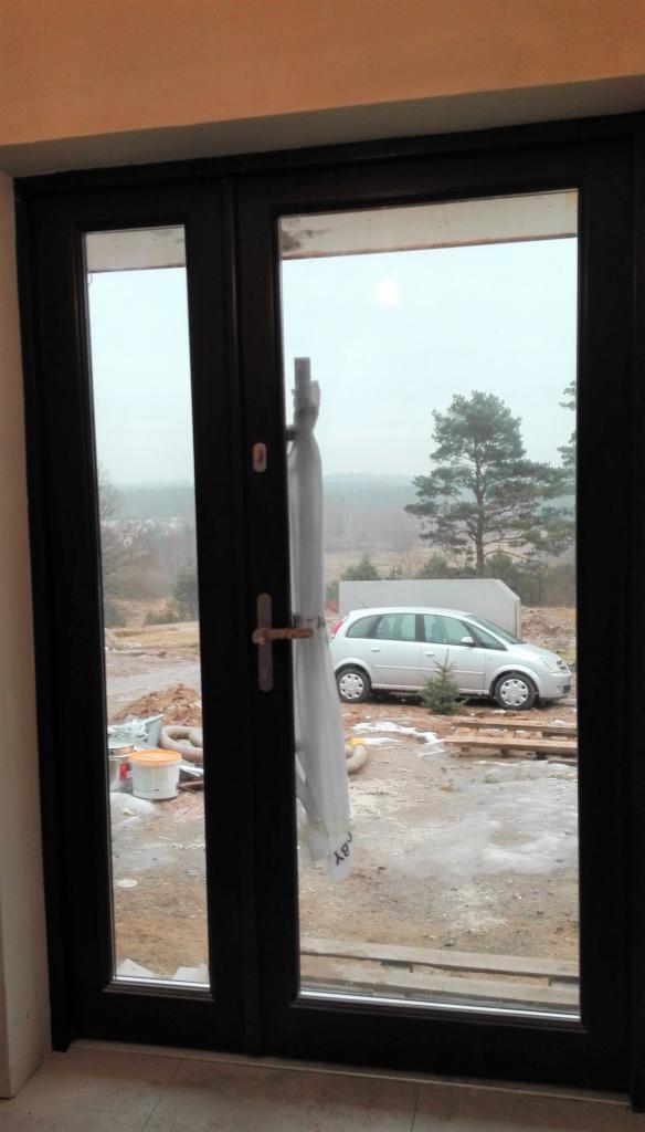 Ciemne, dwuskrzydłowe drzwi balkonowe z widokiem na plac budowy, zaparkowany samochód i drzewa w tle, z widoczną folią ochronną na klamce.