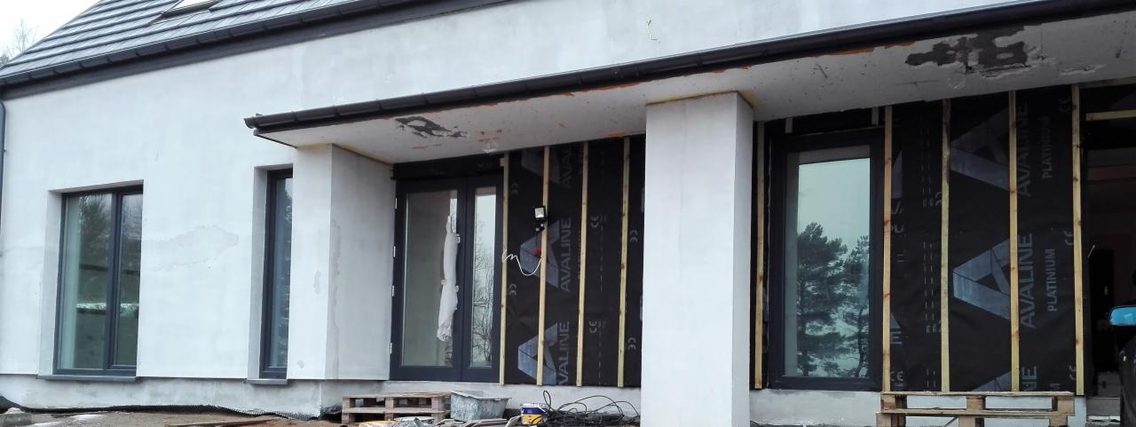 Budowa domu murowanego w trakcie montażu okien i drzwi, widoczne drewniane elementy konstrukcyjne i folia izolacyjna.