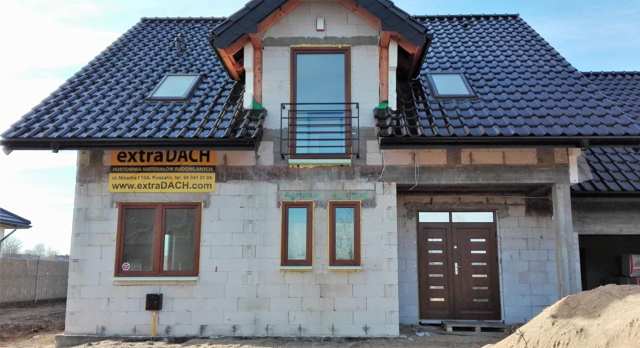 Dom w stanie surowym zamkniętym z brązowymi oknami i drzwiami, ciemna dachówka, widoczny szyld hurtowni materiałów budowlanych.