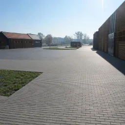 Szeroki chodnik z szarej kostki prowadzący wzdłuż nowoczesnego budynku z elementami drewnianymi i metalowymi, z trawnikiem po lewej stronie i budynkami w tle, oświetlony porannym słońcem.