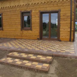 Ułożony taras z dwukolorowej kostki w szachownicę przed drewnianym domem z dekoracyjnymi ramami okiennymi i schodami wejściowymi.