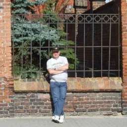 Mężczyzna w dżinsach i koszulce stoi oparty o mur z cegły z metalowym ogrodzeniem, za którym widać iglaki.