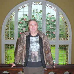 Mężczyzna w kurtce moro stoi na tle dużego, łukowatego okna ze szkła witrażowego przedstawiającego stylizowane liście i kwiaty.