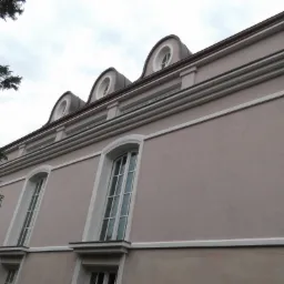 Elewacja budynku w odcieniach beżu z białymi zdobieniami okiennymi i trzema okrągłymi oknami na poddaszu, sfotografowana pod kątem z dołu.