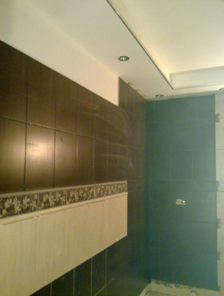 Łazienka w trakcie wykańczania: ciemnobrązowe i kremowe płytki, częściowo ułożone, widoczne taśmy maskujące i niedokończone fugi.