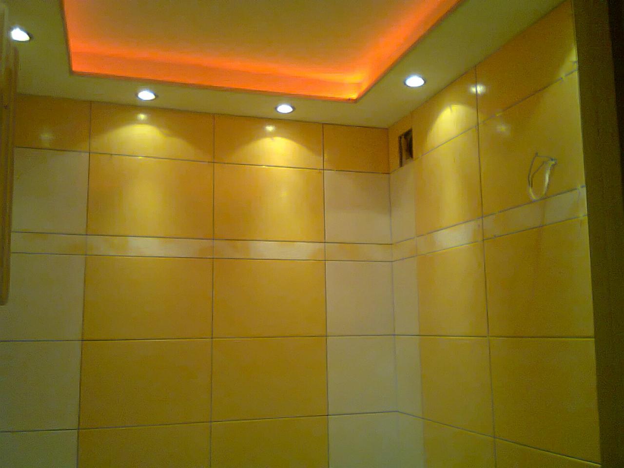 Żółte, błyszczące płytki na ścianach łazienki, oświetlone punktowymi lampami w suficie podwieszanym z pomarańczowym podświetleniem.