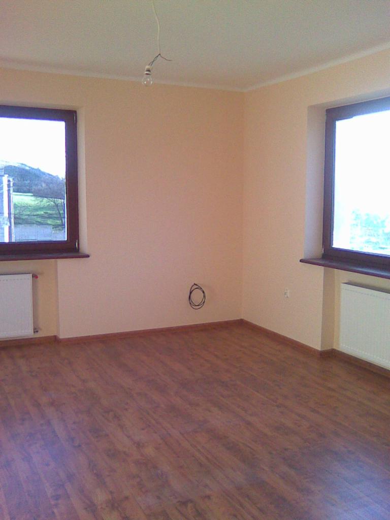 Pomalowane na jasny beż pokój z dwoma oknami z brązowymi ramami, panelami podłogowymi w odcieniach brązu i wiszącą żarówką bez klosza.