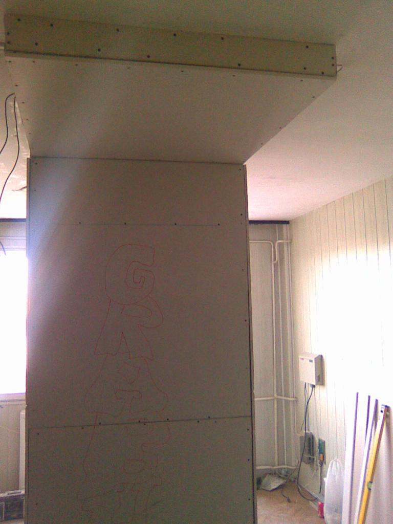 Ściana działowa z płyt gipsowo-kartonowych w trakcie montażu, z widocznym szkicem ołówkiem, mocowana do drewnianej konstrukcji pod sufitem w jasnym wnętrzu.