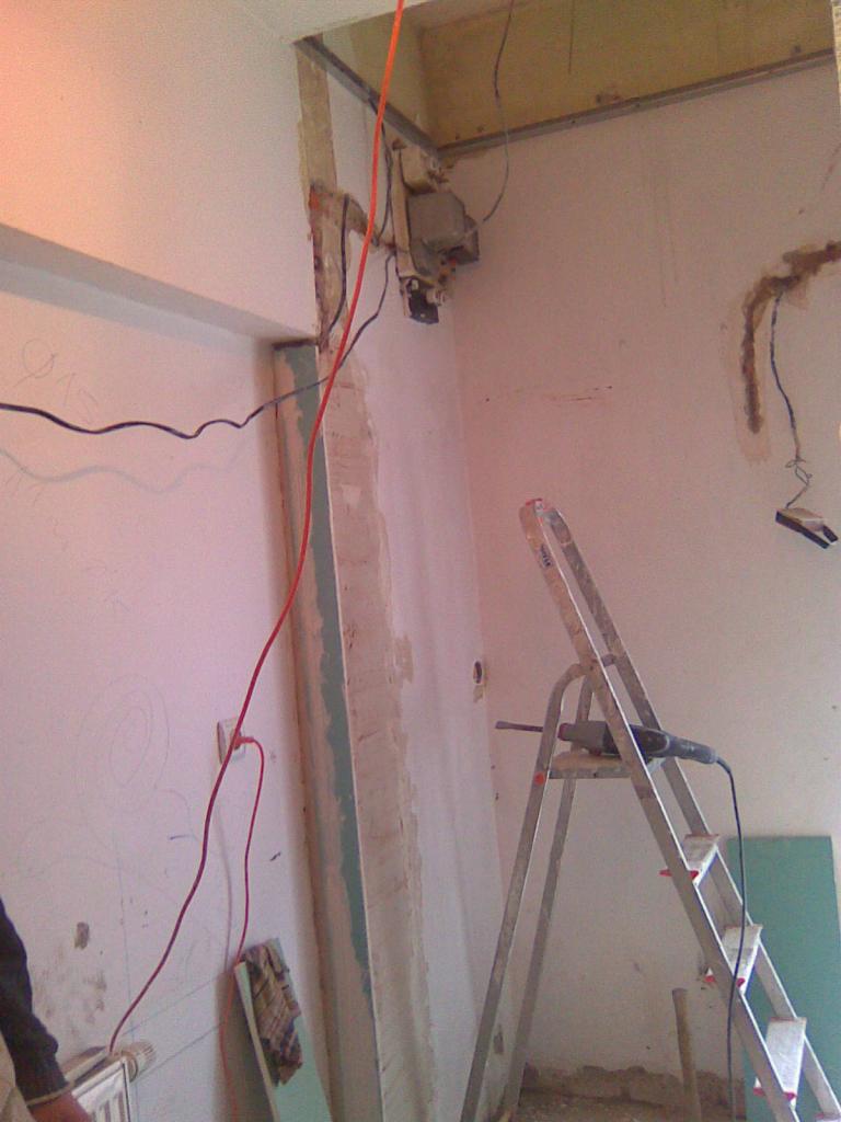 Pokój w trakcie remontu: widoczne przewody elektryczne, drabina z narzędziem, częściowo otynkowana ściana i płyta gipsowa.