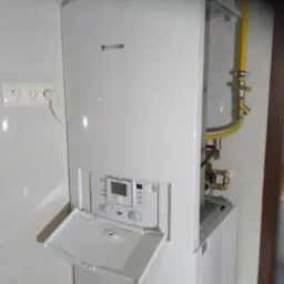 Biały, wiszący piec gazowy z otwartym panelem sterowania, widoczna żółta instalacja gazowa i białe kafelki w tle.