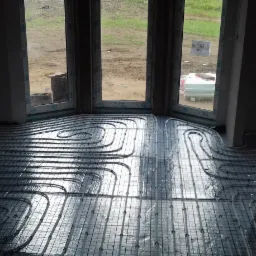 Białe rury ogrzewania podłogowego ułożone w pętle na srebrnej folii izolacyjnej, widok z góry w surowym wnętrzu z oknem wychodzącym na zewnątrz.