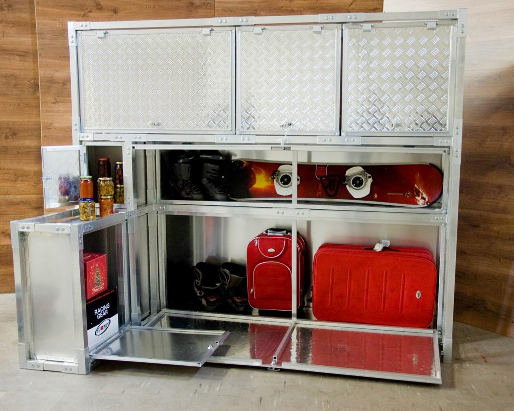 Metalowa szafa garażowa z otwieranymi drzwiczkami i wysuwanymi półkami, zawierająca snowboard, buty narciarskie, walizki, słoiki z przetworami i pudełko z napisem 'RACING GEAR'.