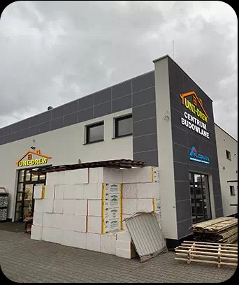 Budynek składu materiałów budowlanych Uni-Drew z logo i stosem bloczków z betonu komórkowego oraz paletami drewna przed wejściem.