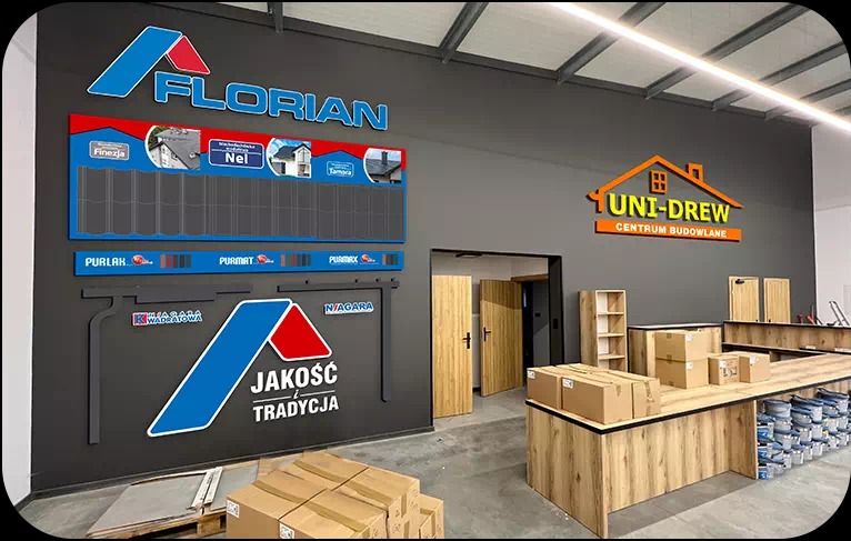Wnętrze składu budowlanego z ekspozycją dachówki ceramicznej Florian i logo Uni-Drew Centrum Budowlane na szarej ścianie.