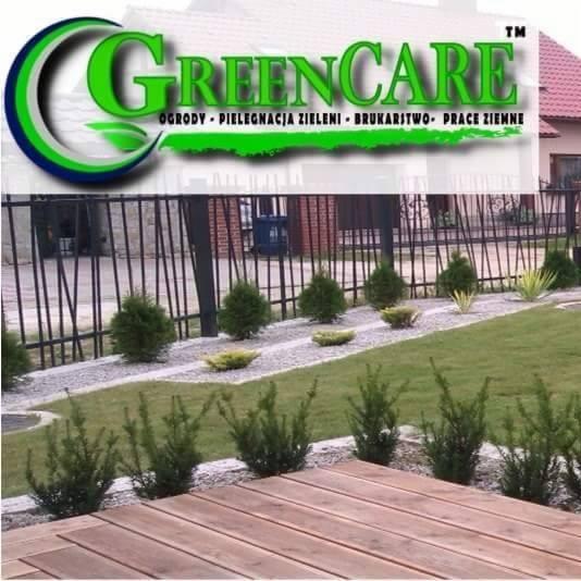 Widok na ogród z drewnianego tarasu, z widocznym logo firmy GreenCare w tle, na zadbanym trawniku nasadzenia z krzewów i kamyków, całość ogrodzona czarnym metalowym płotem.