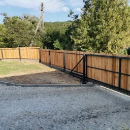 Drewniane ogrodzenie panelowe z przesuwną bramą wjazdową, czarna metalowa konstrukcja, jasne drewno, kostka brukowa na podjeździe, trawnik w tle, słoneczny dzień.