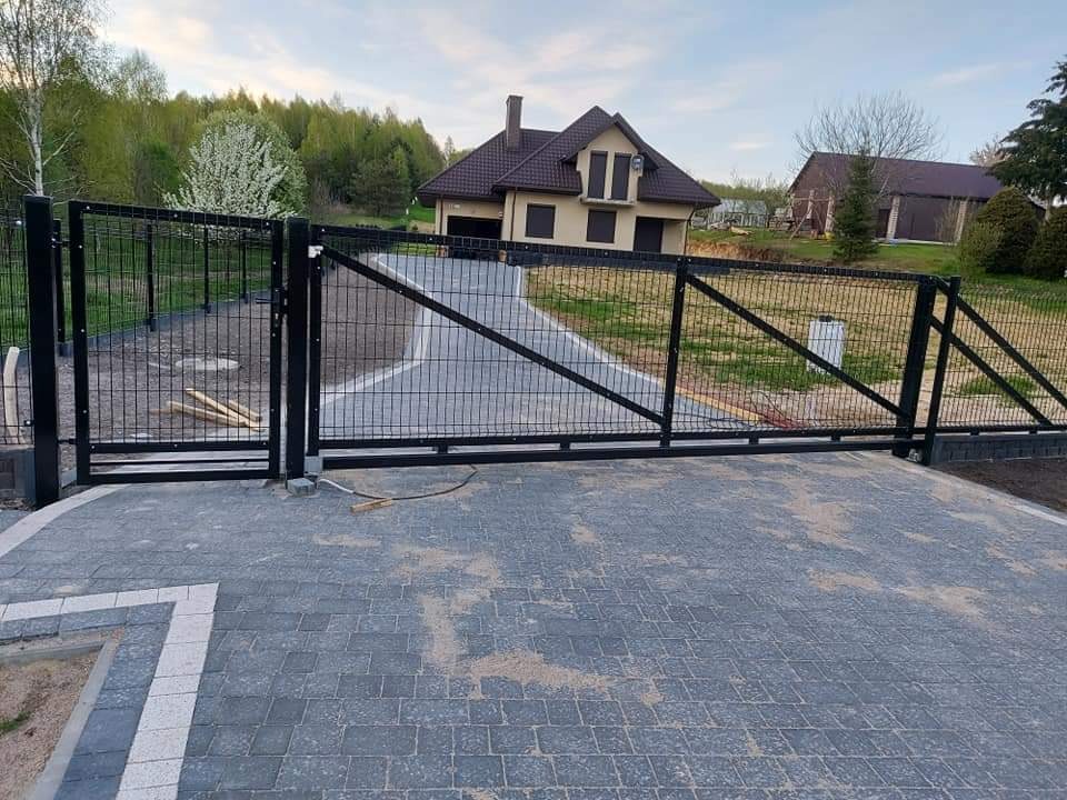 Nowoczesna posesja z przesuwną bramą wjazdową z paneli metalowych, wybrukowany podjazd i dom w tle.
