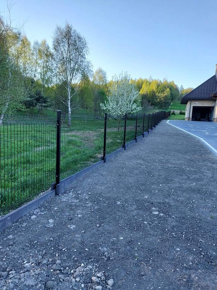 Długi, czarny system ogrodzenia panelowego z podmurówką betonową, widok z perspektywy gruntu, trawnik i drzewa w tle, częściowo wykończony teren.