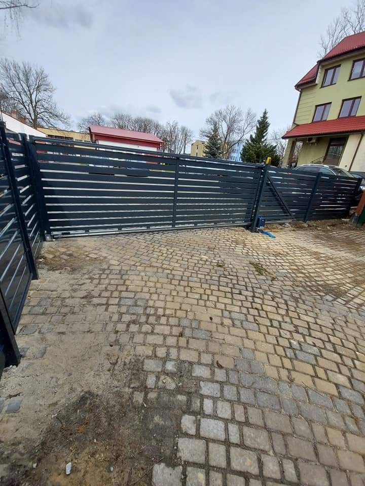 Nowoczesna, pozioma, przesuwna brama wjazdowa z metalowymi panelami na posesji wyłożonej kostką brukową, w tle budynek mieszkalny.