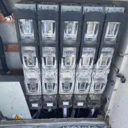 Otwarta skrzynka elektryczna z widocznymi bezpiecznikami APATOR ARS 2 400A, kable i oznaczenie ostrzegawcze przed porażeniem prądem, na tle szarego żwiru.