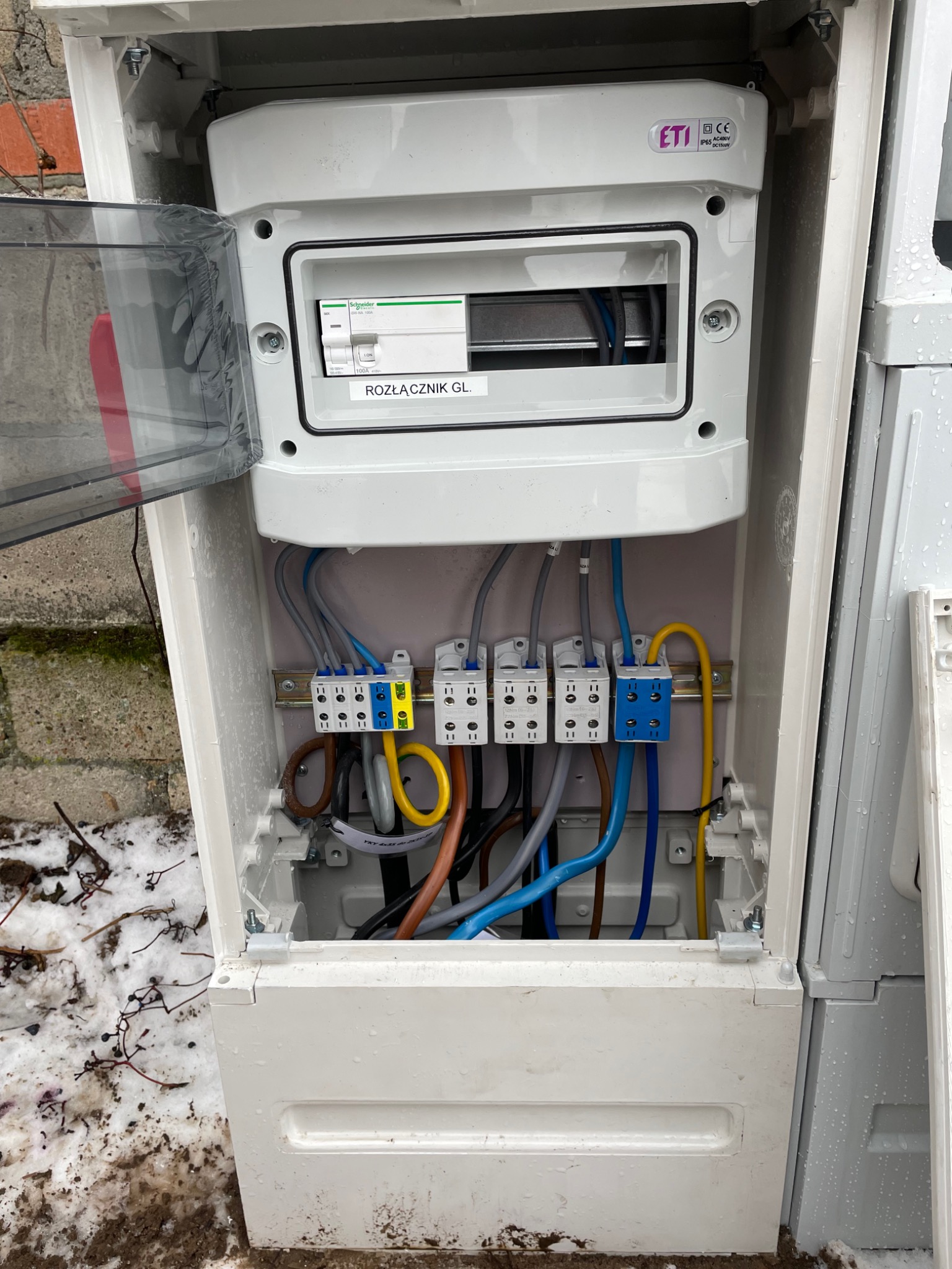 Otwarta, biała skrzynka rozdzielcza z widocznymi bezpiecznikami, rozłącznikiem GL Schneider, listwą zaciskową i uporządkowanymi przewodami w kolorach niebieskim, żółtym i brązowym. Widoczna...