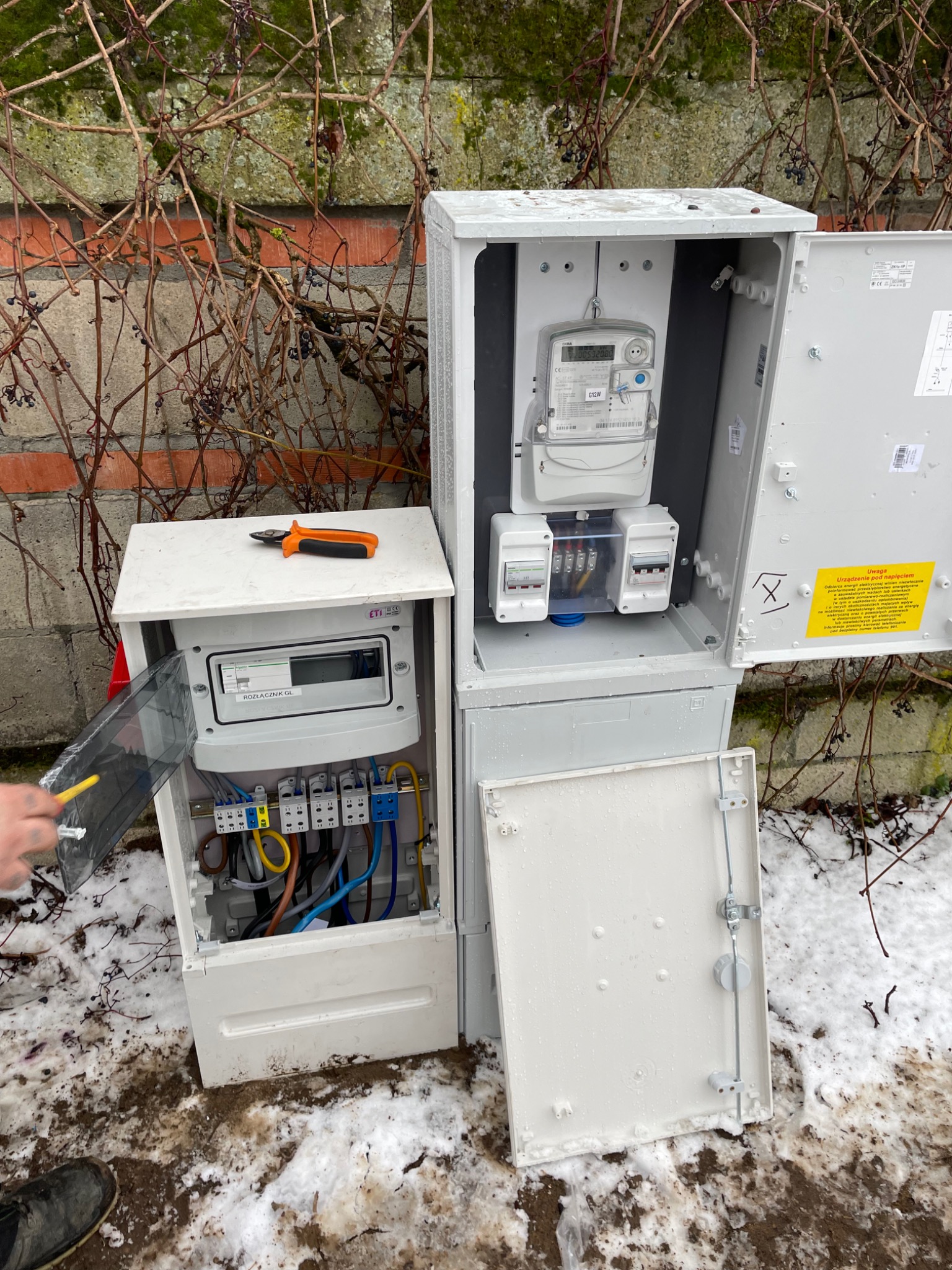 Otwarta skrzynka elektryczna z licznikiem i bezpiecznikami na zewnątrz budynku, z elementami instalacji w trakcie podłączenia, obok leżą obcęgi, a w tle widać ścianę z pnączami.