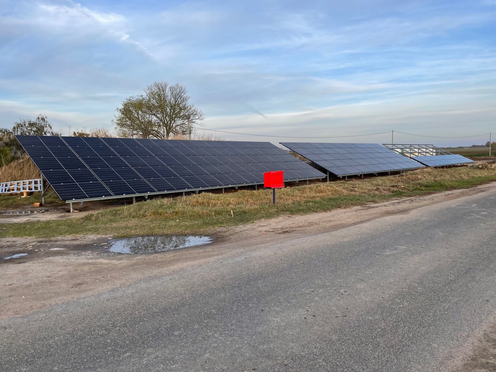 Rząd paneli słonecznych w trakcie instalacji na łące, widoczna metalowa konstrukcja pod panele, obok droga asfaltowa i czerwony słupek.
