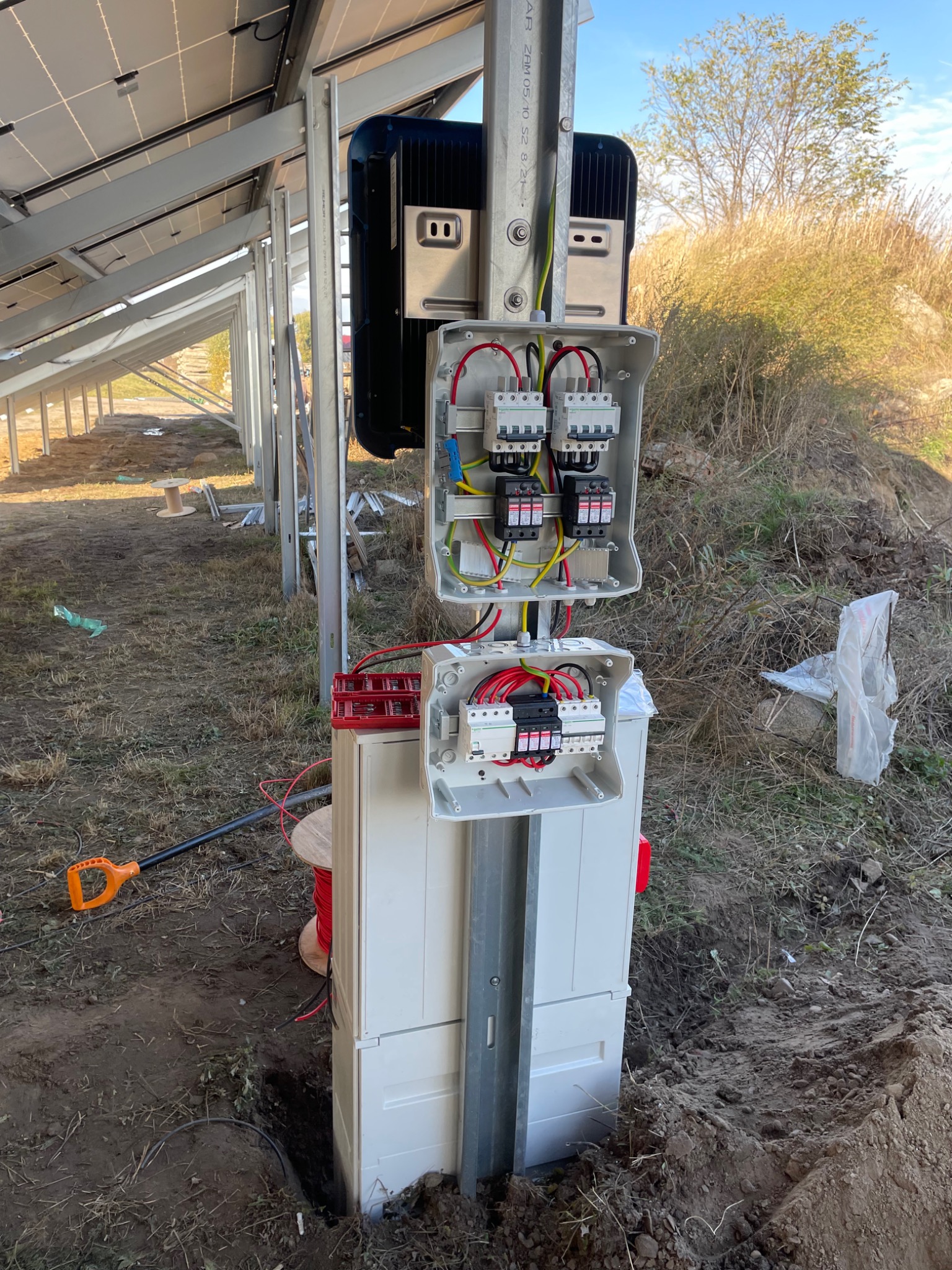 Rozdzielnia elektryczna z otwartymi skrzynkami i widocznymi zabezpieczeniami przepięciowymi, część instalacji fotowoltaicznej w trakcie montażu, na tle paneli słonecznych i niezagospodarowanego...