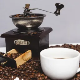 Antyczny młynek do kawy z otwartą szufladką pełną ziaren, otoczony rozsypanymi ziarnami kawy i białą filiżanką z kawą.
