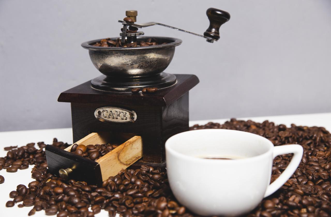 Antyczny młynek do kawy z otwartą szufladką pełną ziaren, otoczony rozsypanymi ziarnami kawy i białą filiżanką z kawą.