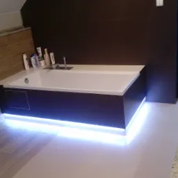 Nowoczesna łazienka z podświetlaną wanną i umywalkami nablatowymi. Brązowe płytki na ścianie, beżowe na podłodze. Widoczne kosmetyki i okno.