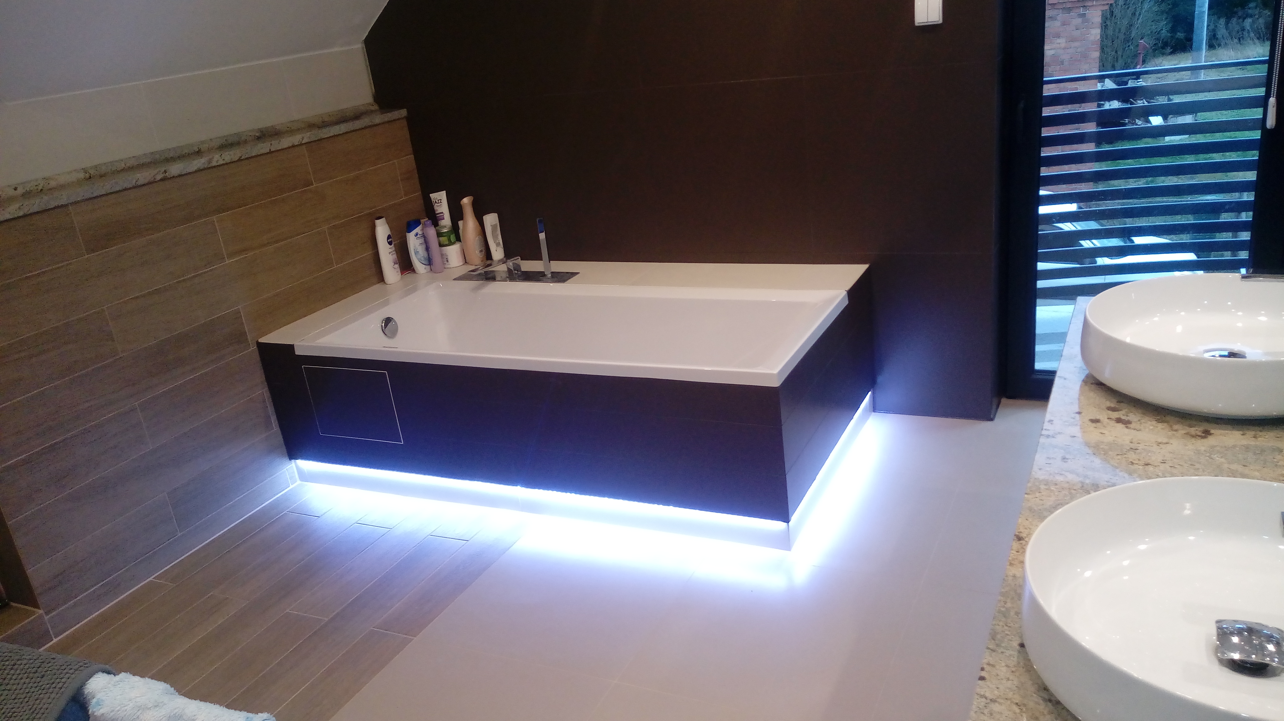 Nowoczesna łazienka z podświetlaną wanną i umywalkami nablatowymi. Brązowe płytki na ścianie, beżowe na podłodze. Widoczne kosmetyki i okno.