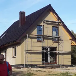 Dom z brązowym dachem w trakcie elewacji, otoczony rusztowaniem. Widoczny fragment osoby i elementy placu budowy. W tle słup wysokiego napięcia.