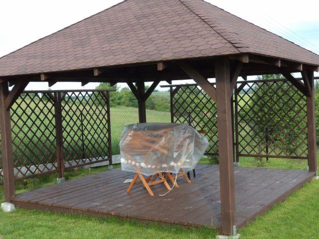 Drewniana altana ogrodowa z brązowym dachem z gontu bitumicznego, obudowana kratownicą, z drewnianą podłogą i meblami przykrytymi folią ochronną.