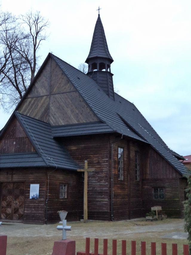 Drewniany kościół z ciemną dachówką, widoczny krzyż na wieżyczce i przed budynkiem, otoczony niskim płotkiem.