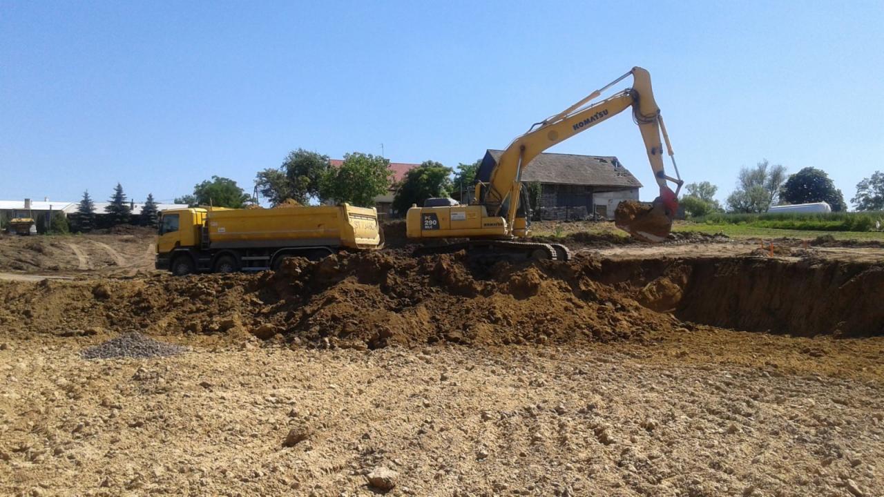 Żółta koparka Komatsu PC290 i wywrotka podczas prac ziemnych na placu budowy w słoneczny dzień. Koparka nabiera ziemię, a w tle widoczny budynek.