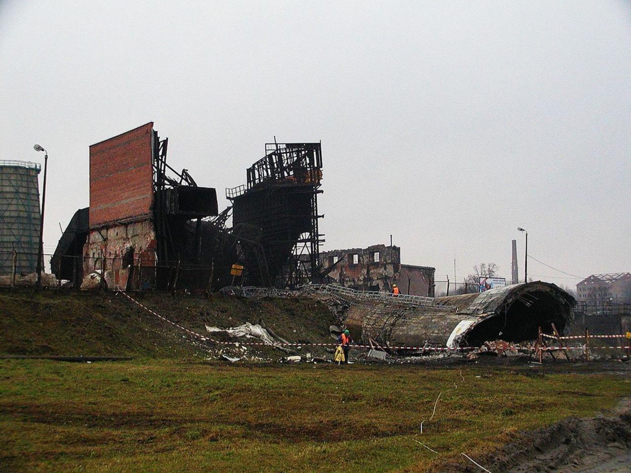 Zdjęcie przedstawia pozostałości po rozbiórce dużego obiektu przemysłowego. Widoczne zniszczone mury, elementy konstrukcji stalowej i gruzowisko na tle pochmurnego nieba.