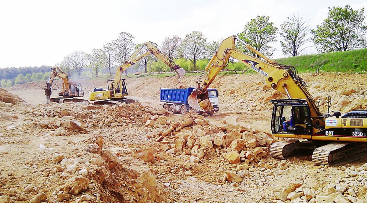 Żółte koparki CAT 325D i ciężarówka podczas prac ziemnych na placu budowy. W tle drzewa i wzgórze.