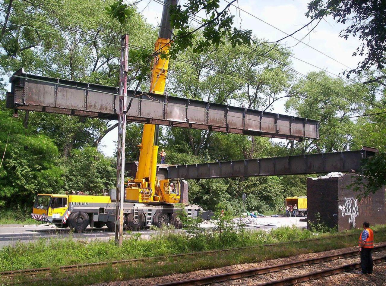Żółty dźwig podnosi stalową konstrukcję mostu nad torami kolejowymi, w tle drzewa i osoby w kamizelkach odblaskowych. Prace rozbiórkowe.