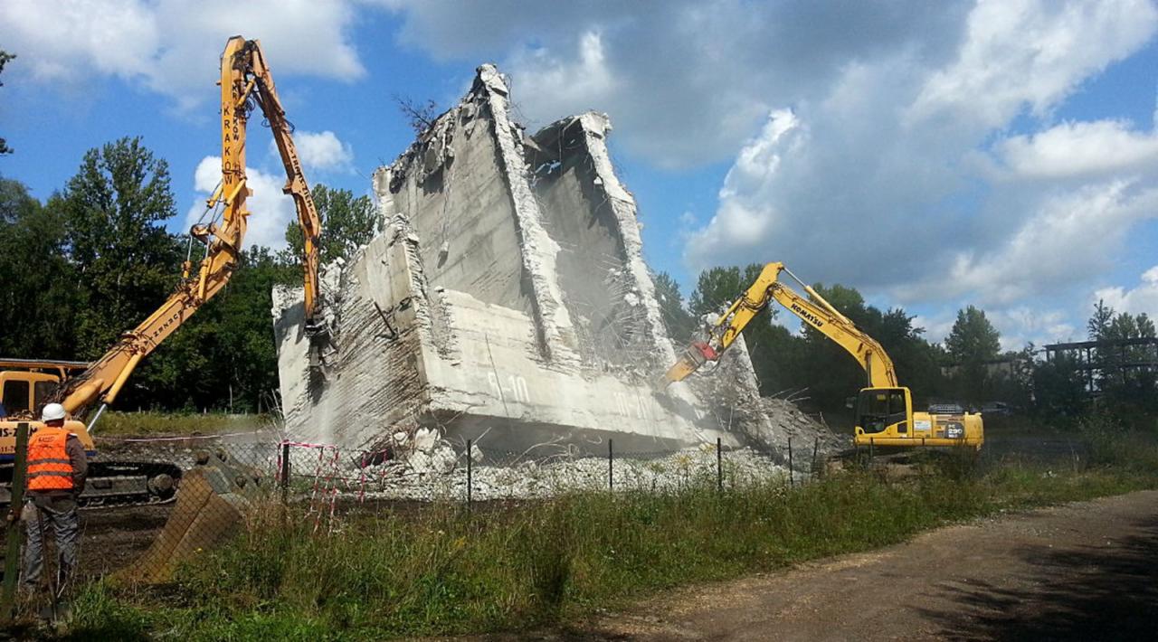Dwa żółte koparki Komatsu kruszą betonową konstrukcję z wystającymi prętami zbrojeniowymi na tle błękitnego nieba z chmurami i zieleni. Na pierwszym planie trawa i siatka.