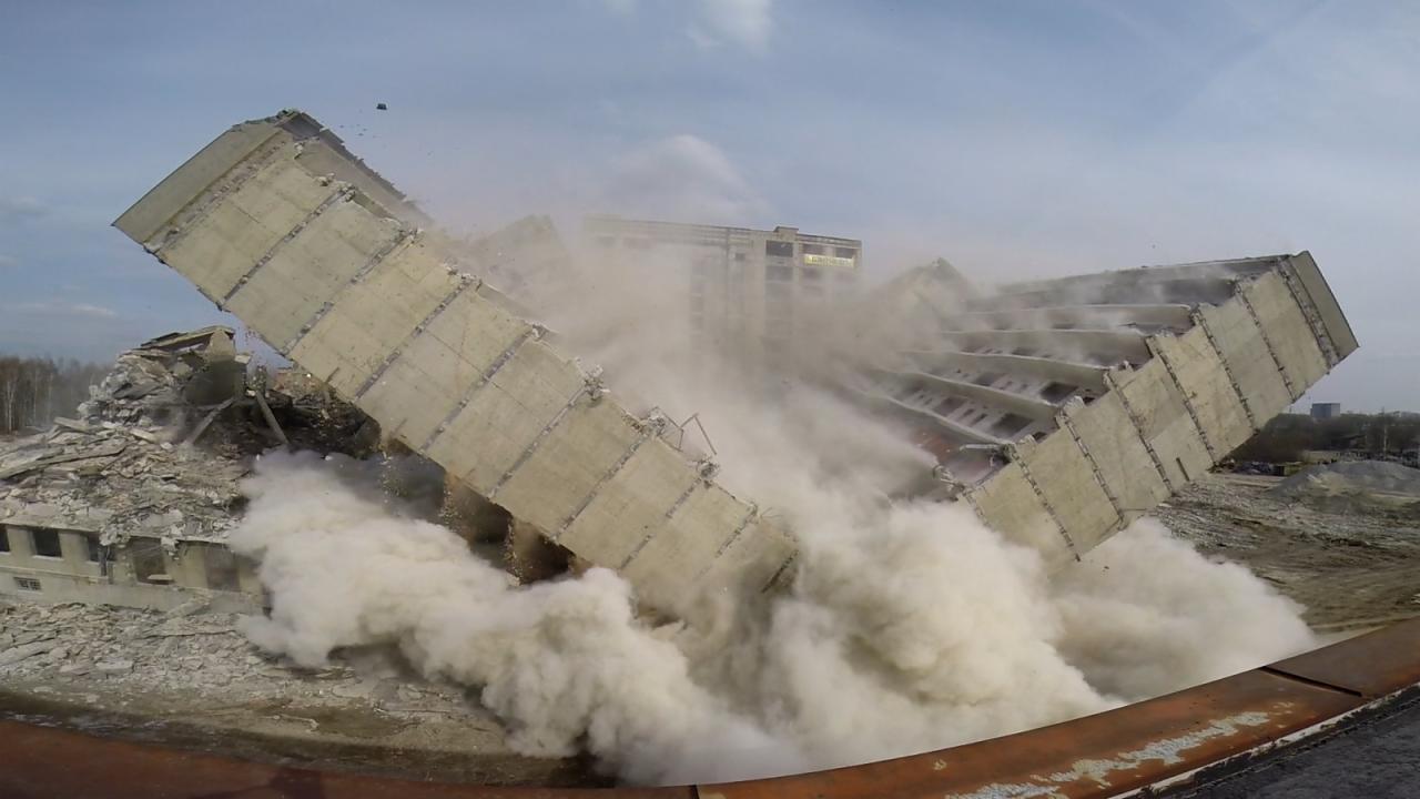 Dynamiczne ujęcie rozpadającego się, betonowego budynku podczas kontrolowanego wyburzenia, z chmurami pyłu unoszącymi się w powietrzu i fragmentami konstrukcji w locie.