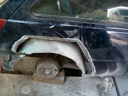 Uszkodzony błotnik samochodu w trakcie naprawy blacharskiej, widoczne ślady spawania i niedoskonałości, podnośnik podtrzymujący element karoserii.