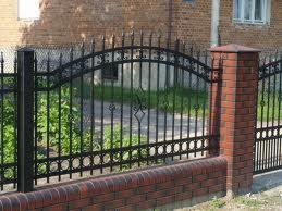 Czarne, metalowe ogrodzenie z dekoracyjnymi elementami, osadzone na niskim murku z czerwonej cegły. W tle widoczny budynek z czerwonej cegły i zieleń.