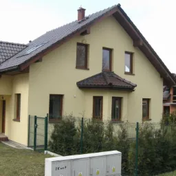 Żółty dom jednorodzinny z brązowymi ramami okiennymi i ciemną dachówką, widoczny z poziomu gruntu za zielonym ogrodzeniem z siatki.