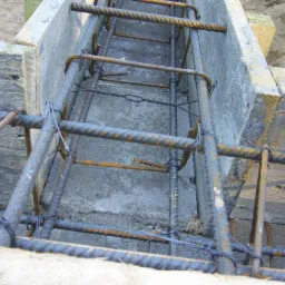 Zbrojenie fundamentu w szalunku z desek, widok z góry. Stalowe pręty połączone drutem, beton wylewany między deskami, przygotowanie do budowy.