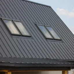 Szary dach z blachy na domu jednorodzinnym, widoczne dwa okna dachowe i fragment wieży kościelnej w tle.