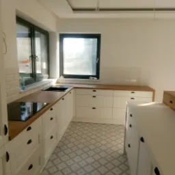 Jasna kuchnia z drewnianym blatem, białymi szafkami z czarnymi uchwytami, wzorzystą podłogą i dwoma oknami, w tym jedno z widocznymi śladami folii ochronnej.