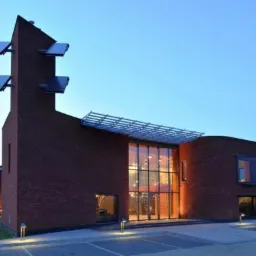 Nowoczesny budynek murowany z czerwonej cegły z dużymi przeszkleniami, nietypową wieżą z panelami słonecznymi i zadaszeniem nad wejściem, oświetlony lampami zewnętrznymi.